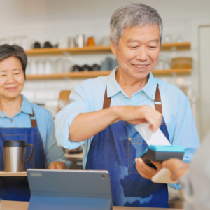 The elderly shop owners smile warmly as they complete a transaction. The customer is using his cellphone to make a tap payment on a digital device, demonstrating the convenient and seamless nature of cashless payment methods in Singapore.