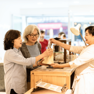 Two joyful elderly women in a boutique, happily receiving their purchase in a paper bag from a smiling shop attendant. This image illustrates the ease and enjoyment of shopping with cashless payment methods for seniors in Singapore.