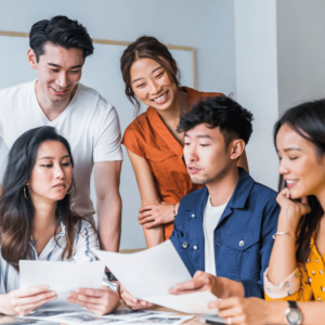 A group of five cheerful young adults, three of whom are seated, engrossed in analyzing printed financial data. Some documents are scattered on the tale, embodying a collaborative and joyful approach to mastering budgeting skills and financial planning in Singapore. This image highlights the potential of teamwork in tackling financial challenges and achieving financial stability.