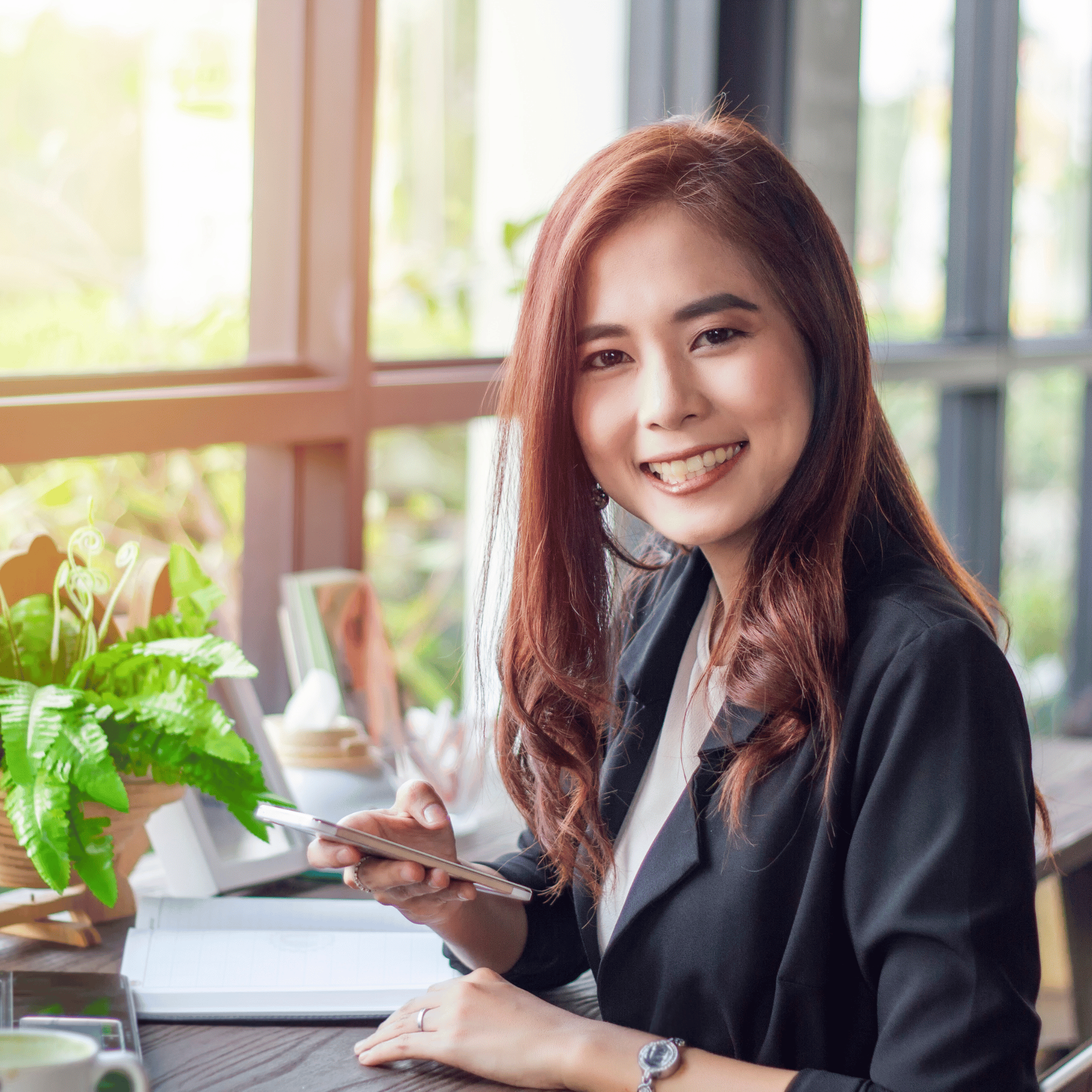 A smiling woman sits at a table in a comfortable office, illustrating the proactive approach to personal finance management. She is holding a cell phone in one hand, representing modern digital methods, while a notebook lies open on the table before her, suggesting the practice of planning and keeping track of expenses.