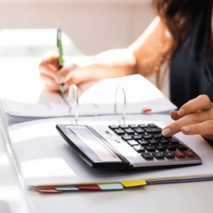 A focused woman sitting at a desk, holding a pen in one hand and a calculator in the other. She is diligently filling out a form. A folder containing relevant documents is also visible on the desk.