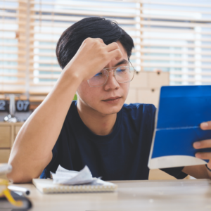 A young Singaporean man looking worried as he sits in his home office, surrounded by bills and scrutinizing his bank book, reflecting the harsh financial realities brought about by the COVID-19 pandemic