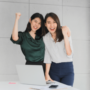 Two enthusiastic millennial women celebrating their financial planning achievements in a vibrant office environment, surrounded by essential tools like a laptop, documents, and a mobile phone, illustrating the positive outcomes of seeking professional financial advice for robust and reliable financial planning.