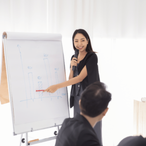 A group of enthusiastic millennials in a tidy conference room, deeply engaged in a discussion about financial literacy in Singapore. A woman, holding a wireless microphone, is energetically explaining a financial chart, embodying the zeal of young Singaporeans to enhance their financial acumen.