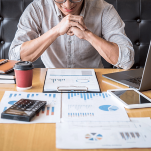 A smiling man, demonstrating a serious yet happy to commit to personal financial planning, is seated at his desk, attentively analyzing graphs for his first investment, with a laptop, and a calculator as his tools.