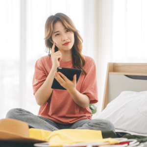 A young woman in a cozy, clean bedroom, deep in thought as she holds a tablet. She's sitting on her bed, surrounded by clothes and a pillow, illustrating the importance of managing finances even in personal spaces. This image underscores the modern approach to financial planning and budgeting in Singapore.