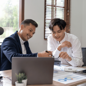 A financial advisor and a young professional engaged in a detailed discussion in a well-lit office. The advisor points to a graph on a document, signifying the sharing of crucial financial insights. The presence of a laptop and other documents filed with charts and graphs on the desk emphasizes the data-driven approach toward setting and achieving financial goals.