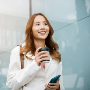 A cheerful young woman strolls along a street in Singapore, coffee in one hand and mobile phone in the other, depicting the positive effects of balanced financial finance on personal well-being.