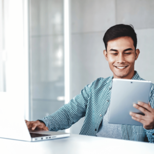 A man in a modern, well-lit home office, engrossed in his tablet smiling, with a laptop nearby. This setting reflects a dynamic approach to personal finance management, utilizing various digital tools for setting financial goals and measuring success in the pursuit of prosperity.