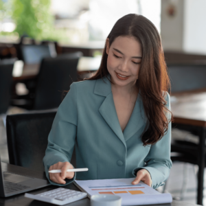 A woman in a cozy cafe setting engrossed in her personal finance planning. She holds a pen, poised above an open notebook, suggesting careful budgeting and expense tracking, while a calculator on the table underscores the importance of precise financial calculations in maintaining a healthy credit history.