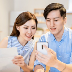 A young, happy couple engaging in early retirement planning; the man is holding a cell phone, while the woman holds a paper, symbolizing their collaborative approach to financial planning.