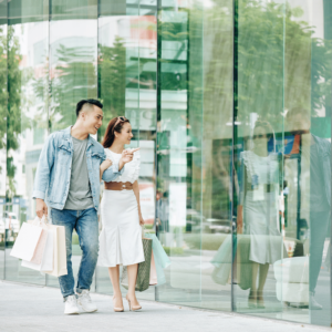 Joyful young couple carrying shopping bags, exploring a bustling shopping district in Singapore. The husband is pointing at something interesting in a store, both are smiling and seem to enjoy their shopping spree. It shows that they are living within their means and have managed their money quite well.