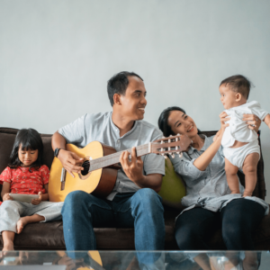 A cheerful family of four spending quality time in their living room, embodying resilience and happiness amidst economic challenges caused by COVID-19, with the father playing the guitar, mother holding a baby, and the older daughter engrossed in her mobile phone.