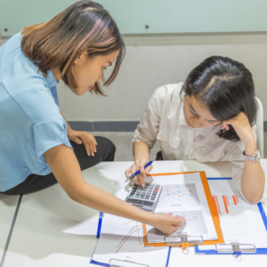 Two young adult women in an office setting, deeply engrossed in analyzing financial data printed on paper. One woman, seated at the desk, is armed with a pen and calculator, underlining the active role young adults in Singapore take in understanding and mastering budgeting skills. This image underscores collaborative learning and discussion in personal finance management.