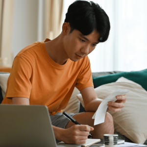 Serious-looking man seated in his living room, intently studying a bill in his left hand, while holding a pen in his right hand, with his laptop open on the desk, symbolizing personal finance management.