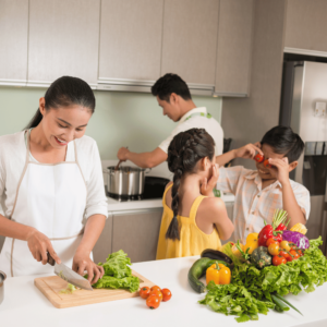 A content Singaporean family of four in their kitchen, illustrating financial stability. The mother chops vegetables while the father cooks, and the two children play happily beside her. This image underscores the theme of adhering to budget plans and achieving financial goals, enabling a wholesome and secure home environment.