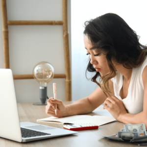 A focused young woman in a clean, modern home office, preparing to write on a notepad with a pen. Beside her are some dollar bills, indicating budgeting tasks, and an open laptop suggests digital tools aiding in financial planning. This image embodies the process of mastering budgeting skills for effective salary management in Singapore.