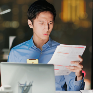 A man is seated in a dimly lit room, looking at a document with a puzzled expression while a computer sits idle on his desk. This image highlights the complexities and challenges that one may face in setting financial goals and measuring success, emphasizing the importance of effective planning and guidance.