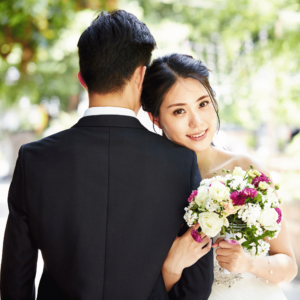 Joyous wedding photo of a Singaporean couple, with the bride holding a bouquet of flowers and her husband's arm, looking happily at the camera while the husband's back is turned towards the camera.